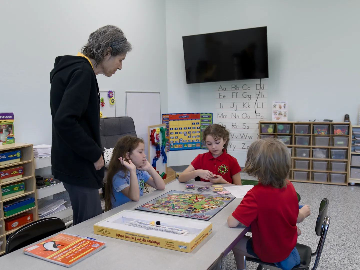 A teacher stands beside three students playing a board game at a table.