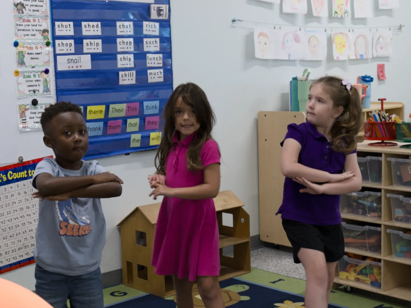 Three elementary students stand in front of a classroom word wall.