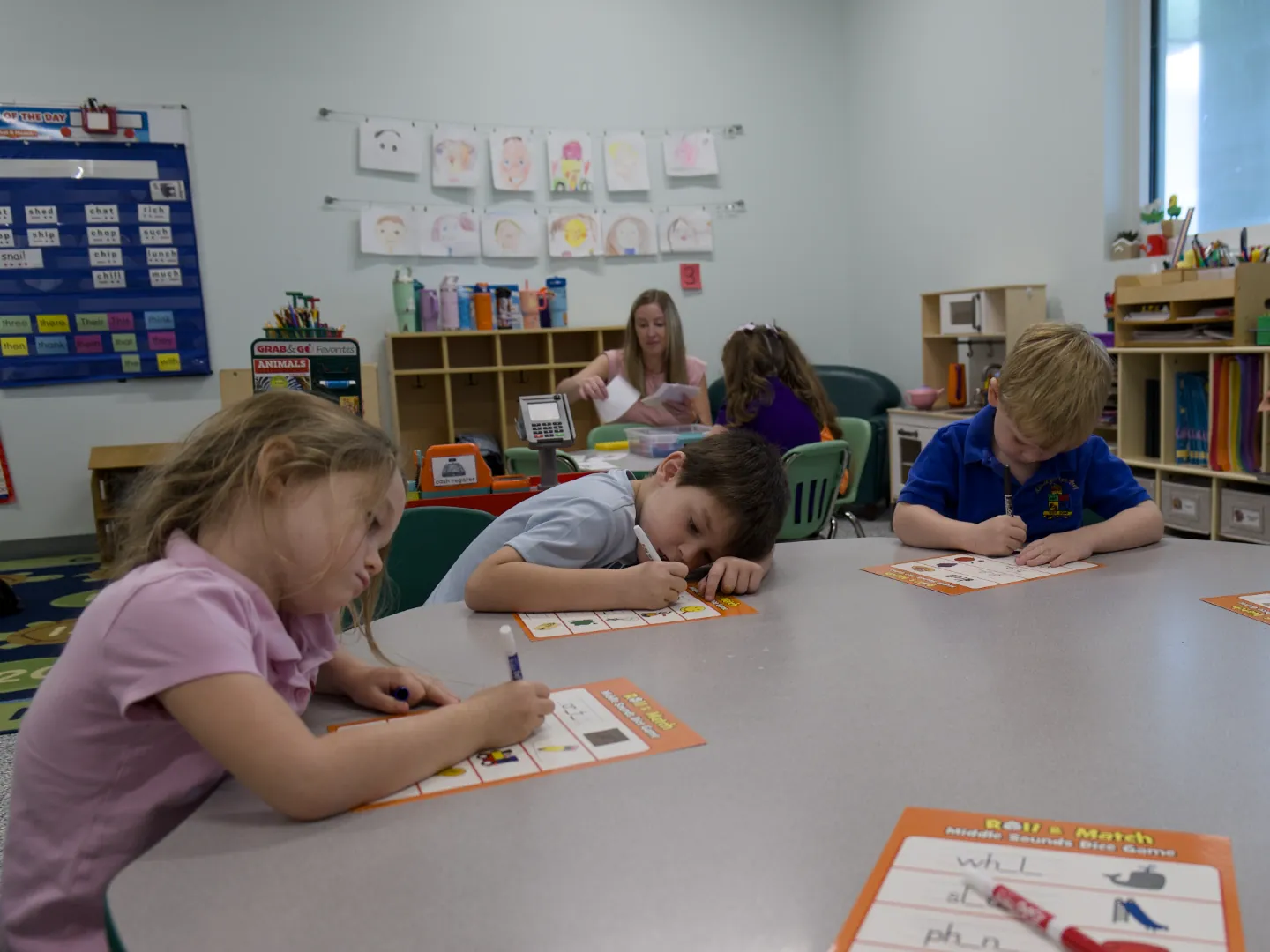 Kindergarten students write at a shared table during a phonics activity.