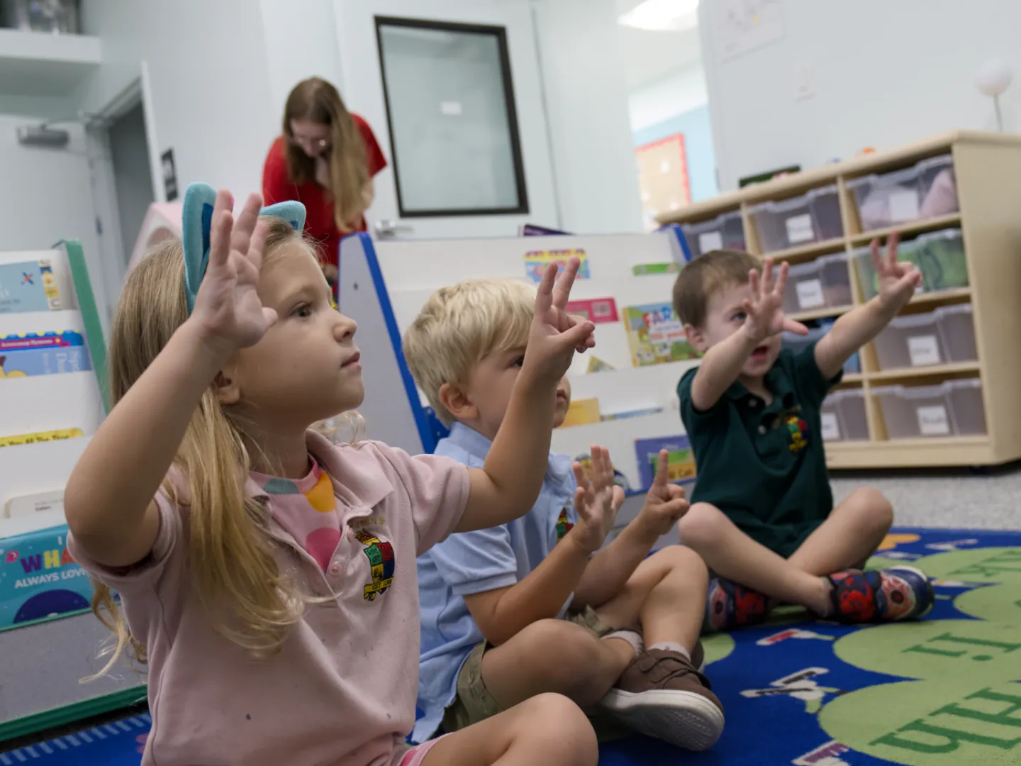 Preschoolers sit on a classroom rug with their hands raised during a counting song.