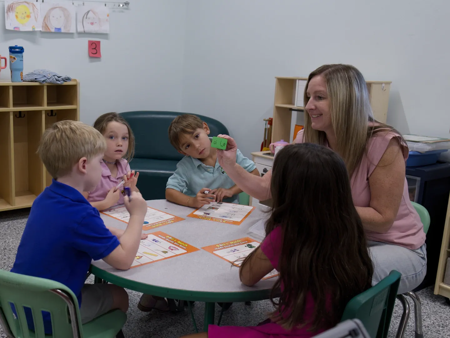 A teacher holds up a letter tile for four children seated around a small table.